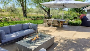Een buitenterras met een vuurkuil, een bank, een tafel met stoelen en een parasol.
