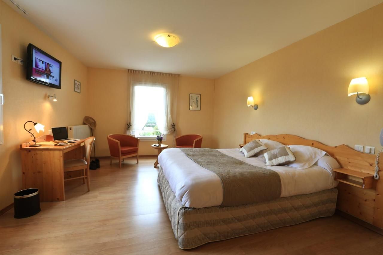 A hotel room with a wooden bed, a desk with a computer, two chairs, a TV, and a window with curtains.