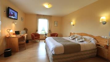 A hotel room with a wooden bed, a desk with a computer, two chairs, a TV, and a window with curtains.