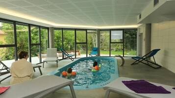 An indoor swimming pool with clear glass walls, lounge chairs, and a ceiling with a grid pattern.