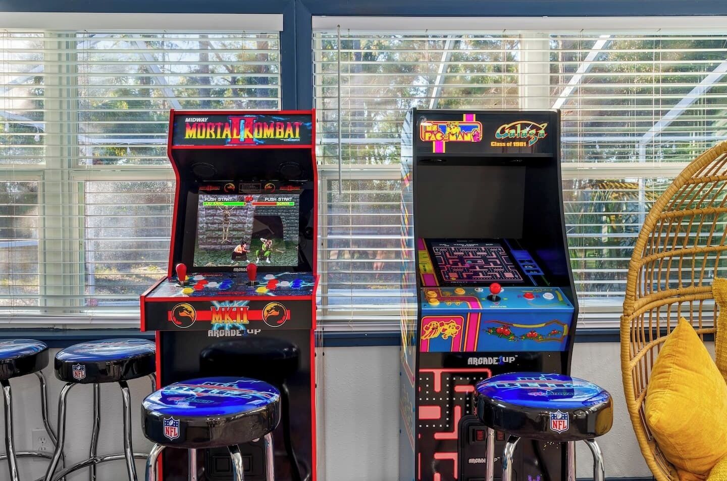 Dos máquinas de videojuegos, una con la etiqueta 'Mortal Kombat' y la otra 'Pac-Man', situadas junto a una ventana con persianas.