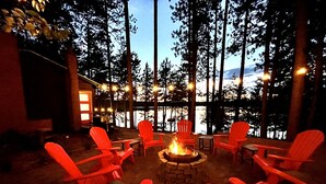 A cozy outdoor seating area with red Adirondack chairs arranged around a central fire pit, surrounded by tall trees and a view of a body of water. - Dollar Lake Retreat: Super Clean, Chef's Kitchen, Hot Tub + Amazing Sunsets (Eagle River)