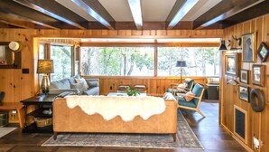 A cozy cabin interior with a sofa, chairs, a dining table, and a large window offering a view of trees. - Dollar Lake Retreat: Super Clean, Chef's Kitchen, Hot Tub + Amazing Sunsets (Eagle River)