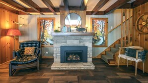 A cozy living room with a stone fireplace, leather armchair, and a staircase with wooden railings. - Dollar Lake Retreat: Super Clean, Chef's Kitchen, Hot Tub + Amazing Sunsets (Eagle River)