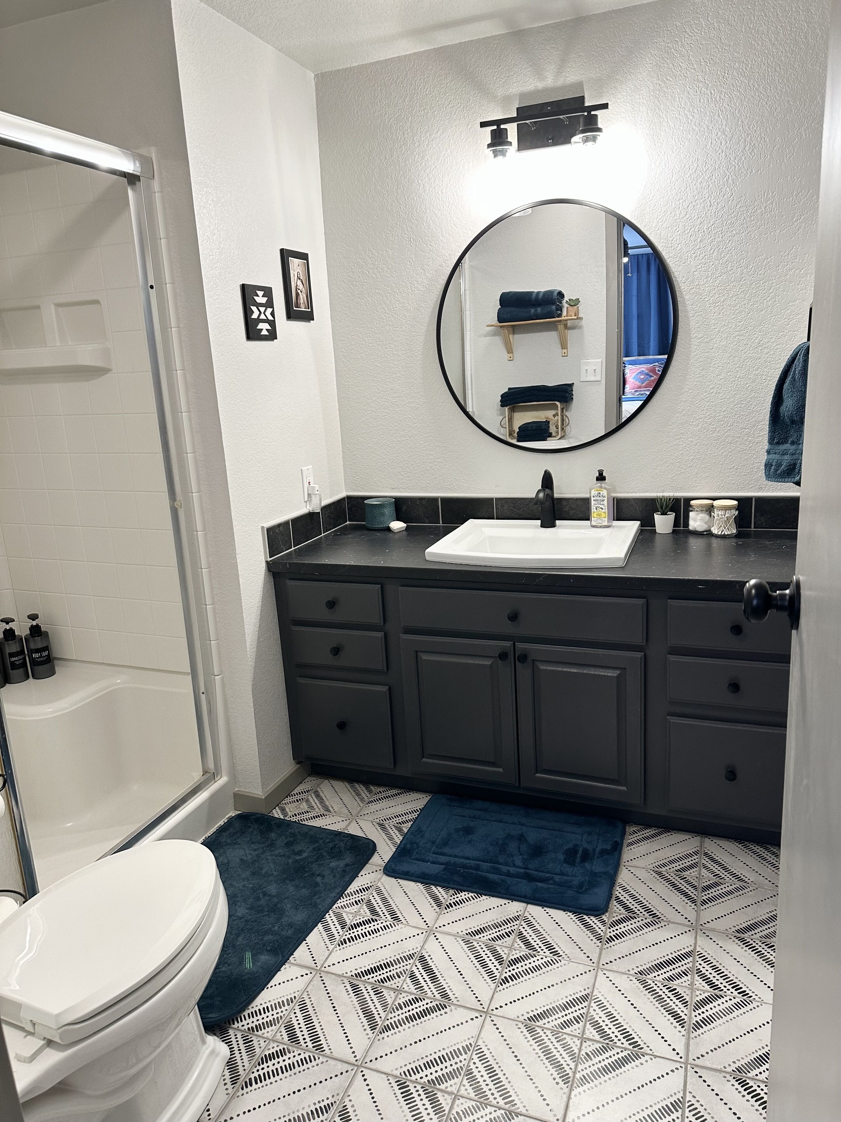 A bathroom with a white toilet, a bathtub, a dark gray vanity with a round mirror, and a patterned floor.