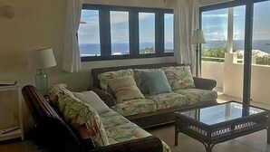 Een woonkamer met een bank, fauteuil en salontafel, grote ramen met uitzicht en een balkon.