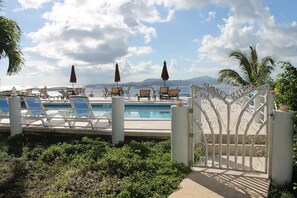 A pool area with lounge chairs and umbrellas, a gated entrance, and a view of the sea and mountains. - Sunset Ridge Villas Unit D (St. John)