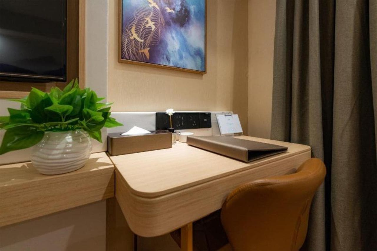 A hotel room with a desk, a laptop, a plant, a painting, and a curtain.