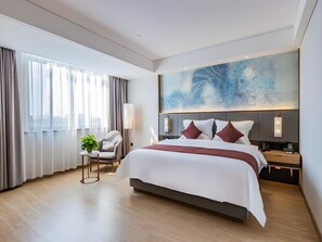 A modern hotel room with a large bed, a desk, and a chair. There is a window with curtains, a painting on the wall, and a plant on a side table.