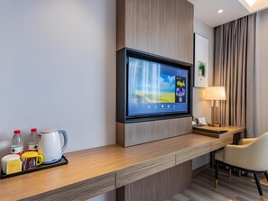 A modern hotel room with a flat-screen TV mounted on a wooden wall, a desk with a lamp, a chair, and a tray with a kettle, cups, and bottles. - Gya Jiaxing Haiyan County Changan Road Lishi Plaza Hotel (Jiaxing)