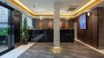 A hotel reception area with a marble reception desk, a large potted plant, and a television screen on the wall.