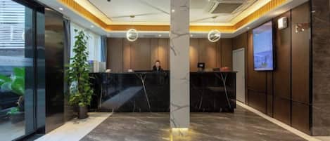 Zona de recepción de un hotel con un mostrador de mármol, una planta grande en maceta y una pantalla de televisión en la pared.
