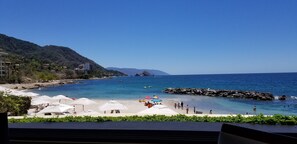 Relax on a well-maintained beach with umbrellas, surrounded by lush greenery and a clear blue sea.