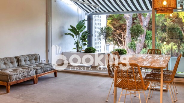 A modern living room with a sofa, dining table, and chairs. There is a large window with a view of greenery and buildings.