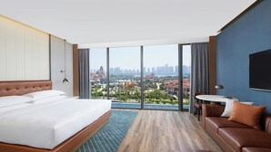 Ein Hotelzimmer mit einem großen Bett, einer Couch, einem kleinen Tisch und einem Fernseher. Das Zimmer verfügt über große Fenster mit Blick auf die Stadt.