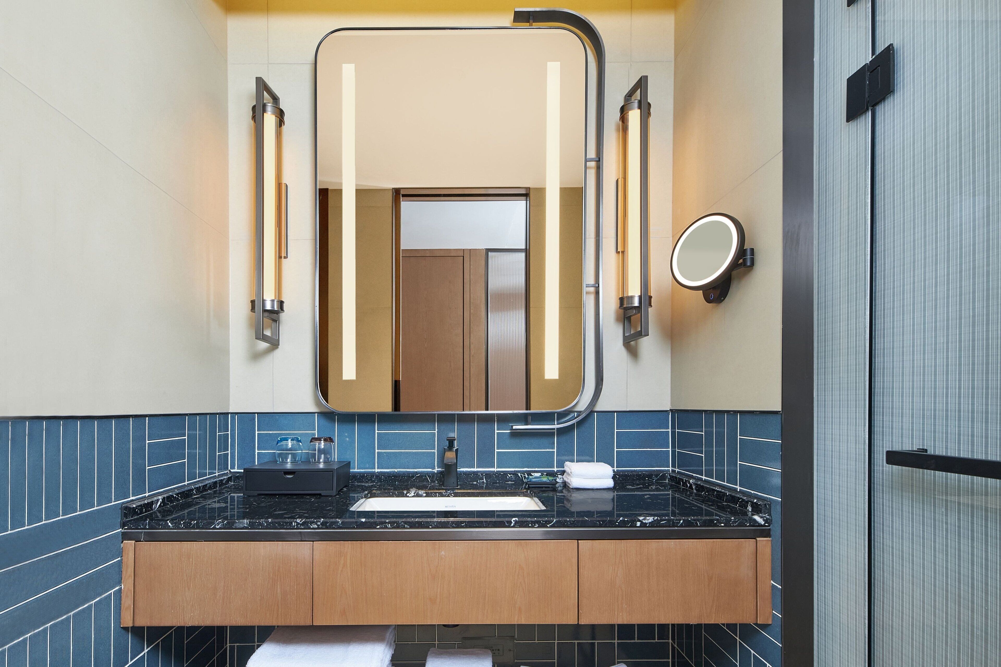 A modern bathroom with a black marble sink, wooden cabinets, and a large mirror with integrated lighting.