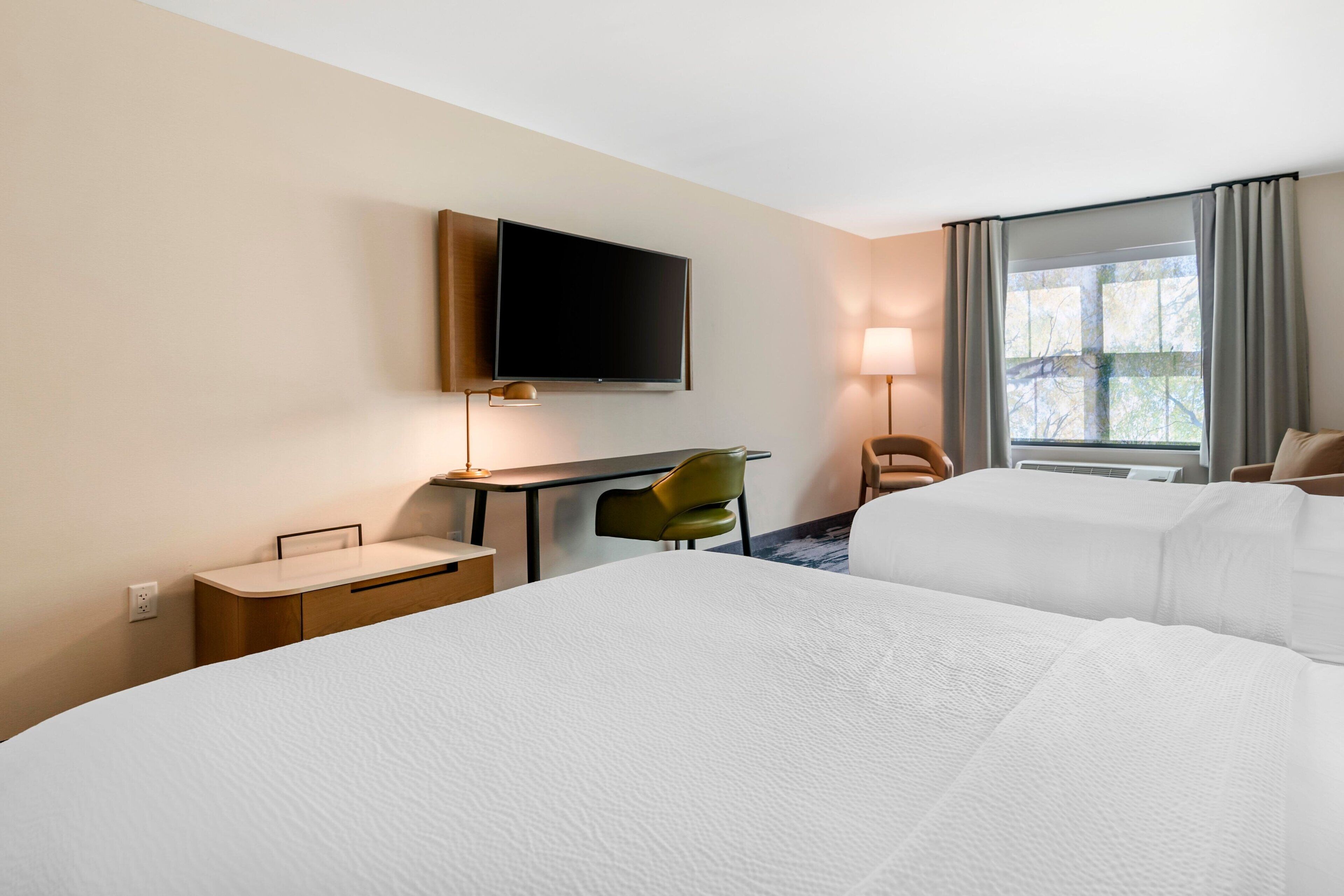 Habitación de hotel con dos camas, un escritorio con silla, televisión montada en la pared y ventana con cortinas.