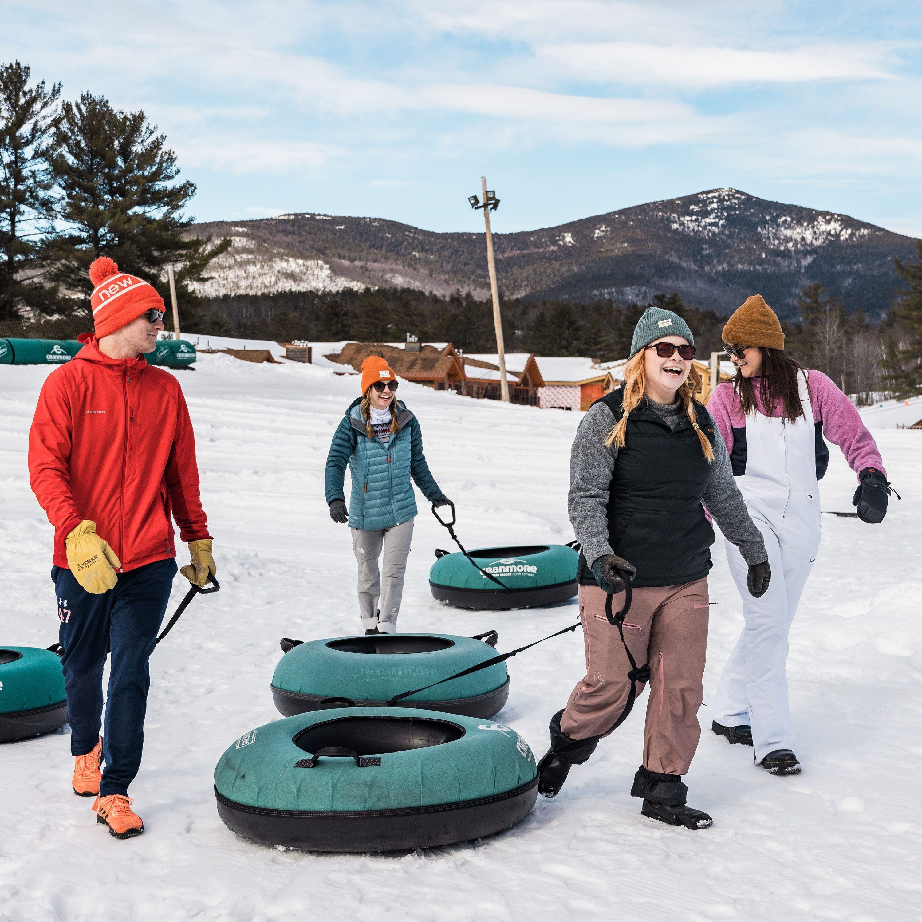 A group of people in winter gear, preparing to use snow tubes on a snowy landscape with mountains in the background.