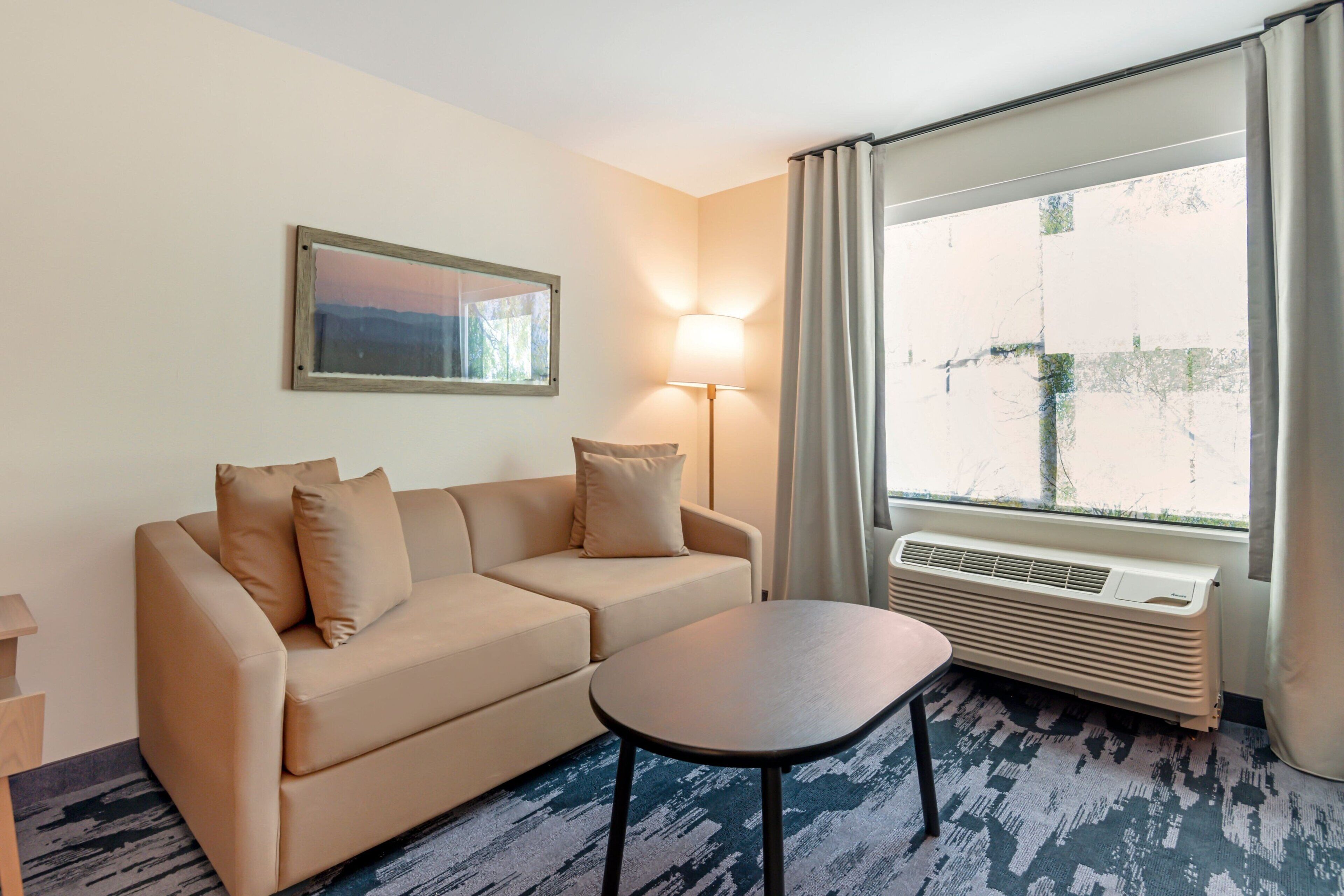 A hotel room with a beige sofa, a small table, a lamp, and a large window with curtains.