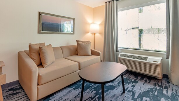 A hotel room with a beige sofa, a small table, a lamp, and a large window with curtains. - Fairfield Inn & Suites By Marriott North Conway (North Conway)