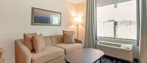 A hotel room with a beige sofa, a small table, a lamp, and a large window with curtains.