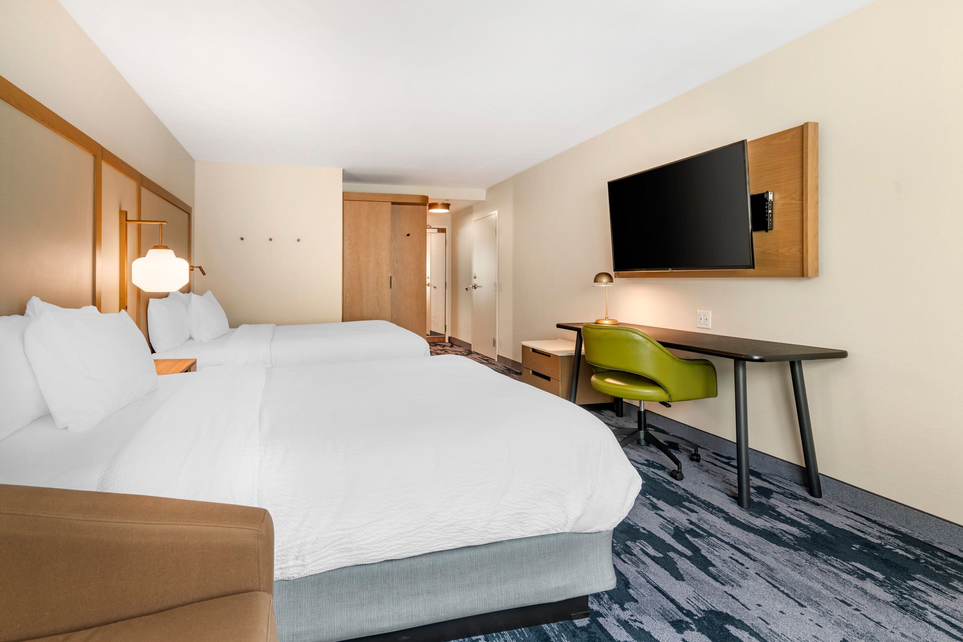 A hotel room with two beds, a desk, a chair, a TV mounted on the wall, and a door leading to another room.