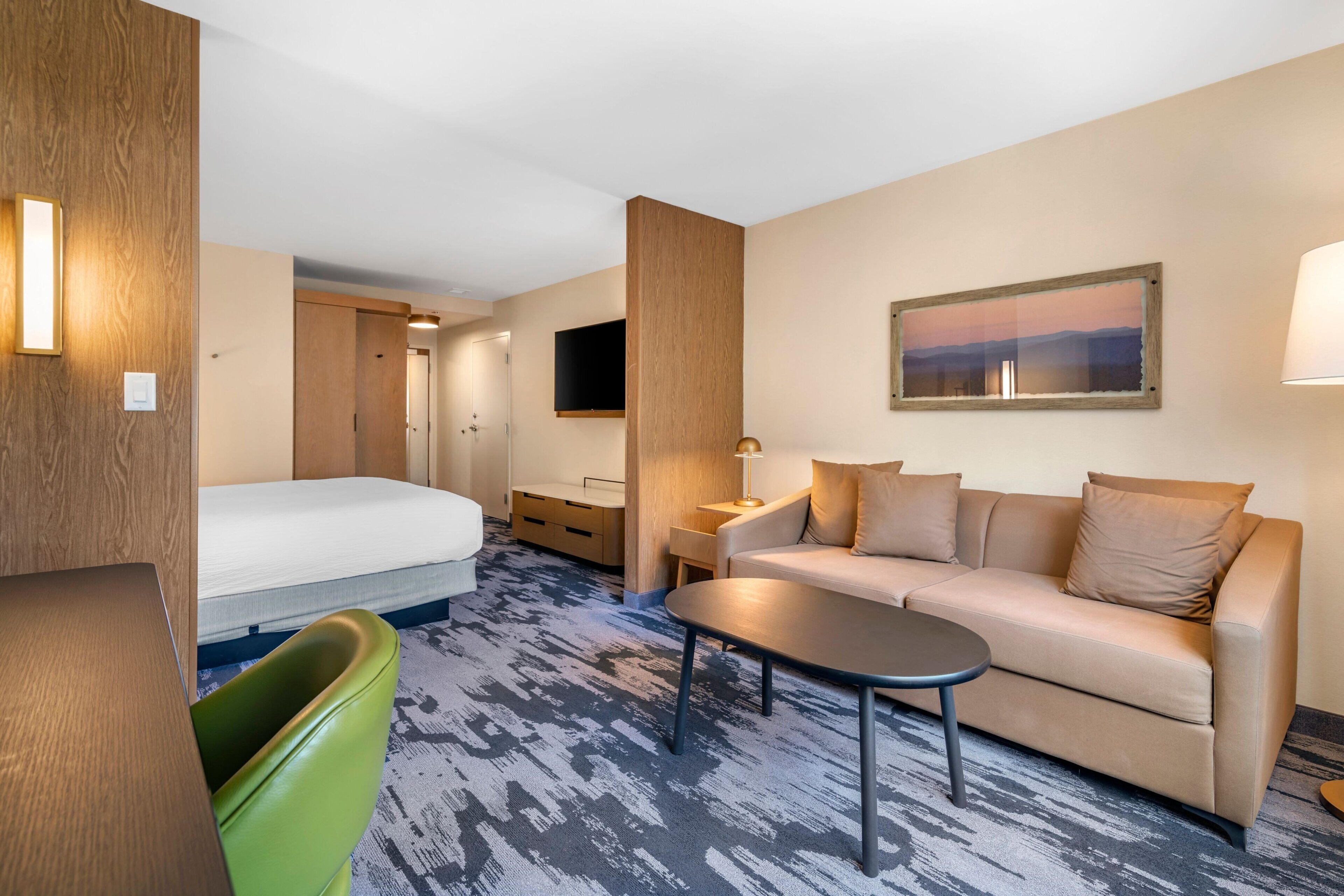 A hotel room with a bed, a sofa, a coffee table, a TV, and a painting on the wall.