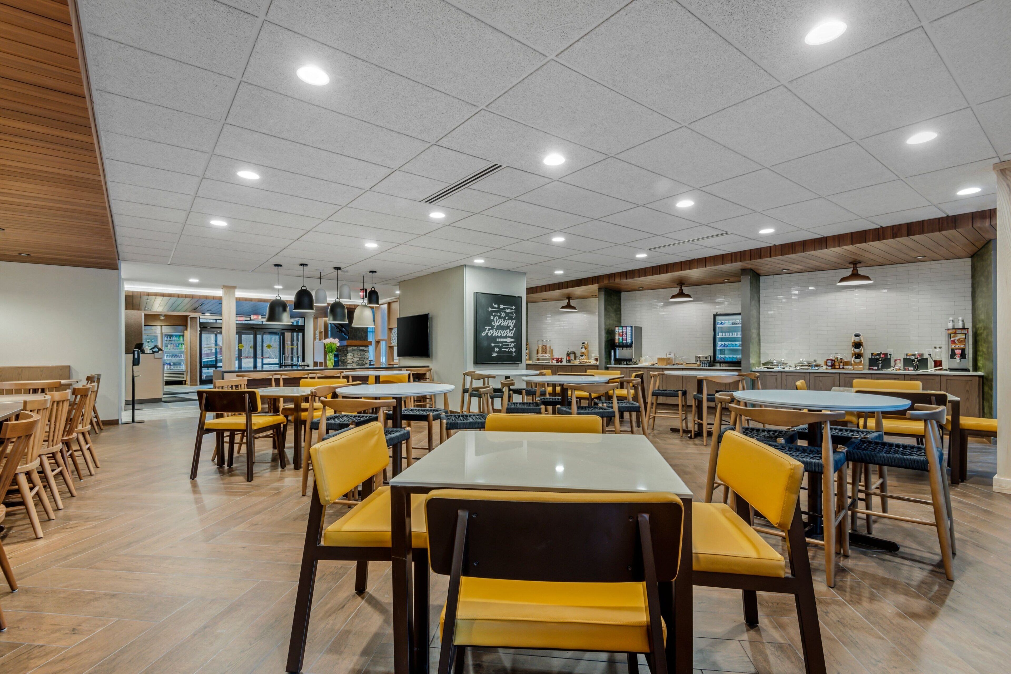A spacious indoor dining area with multiple tables and chairs, a kitchen area in the background, and a chalkboard with writing on the wall.
