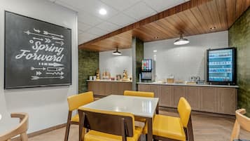 Un restaurante con una mesa blanca y sillas amarillas, una pizarra con mensajes motivadores y un televisor sobre el mostrador.