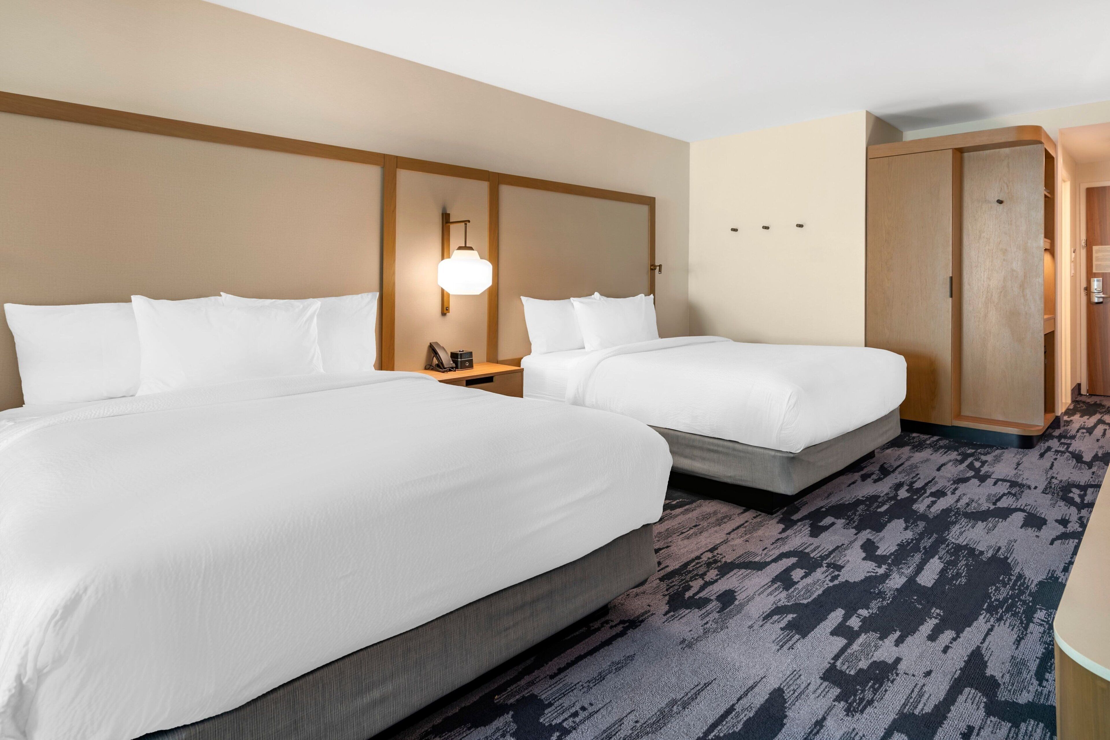 A hotel room with two beds, a wooden wardrobe, a nightstand with a lamp, and a patterned carpet.