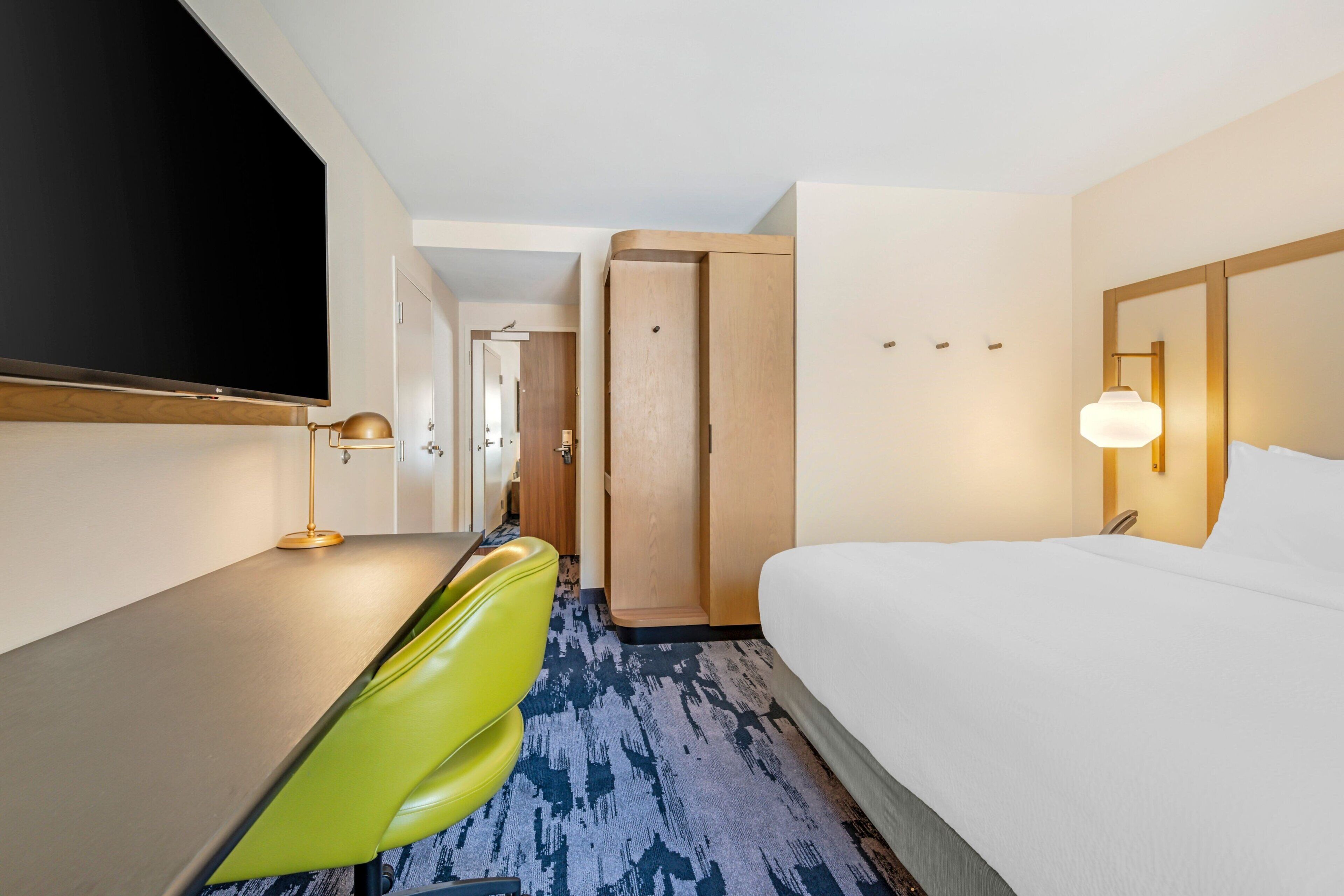 A hotel room with a bed, a desk, a chair, a television, and a door.