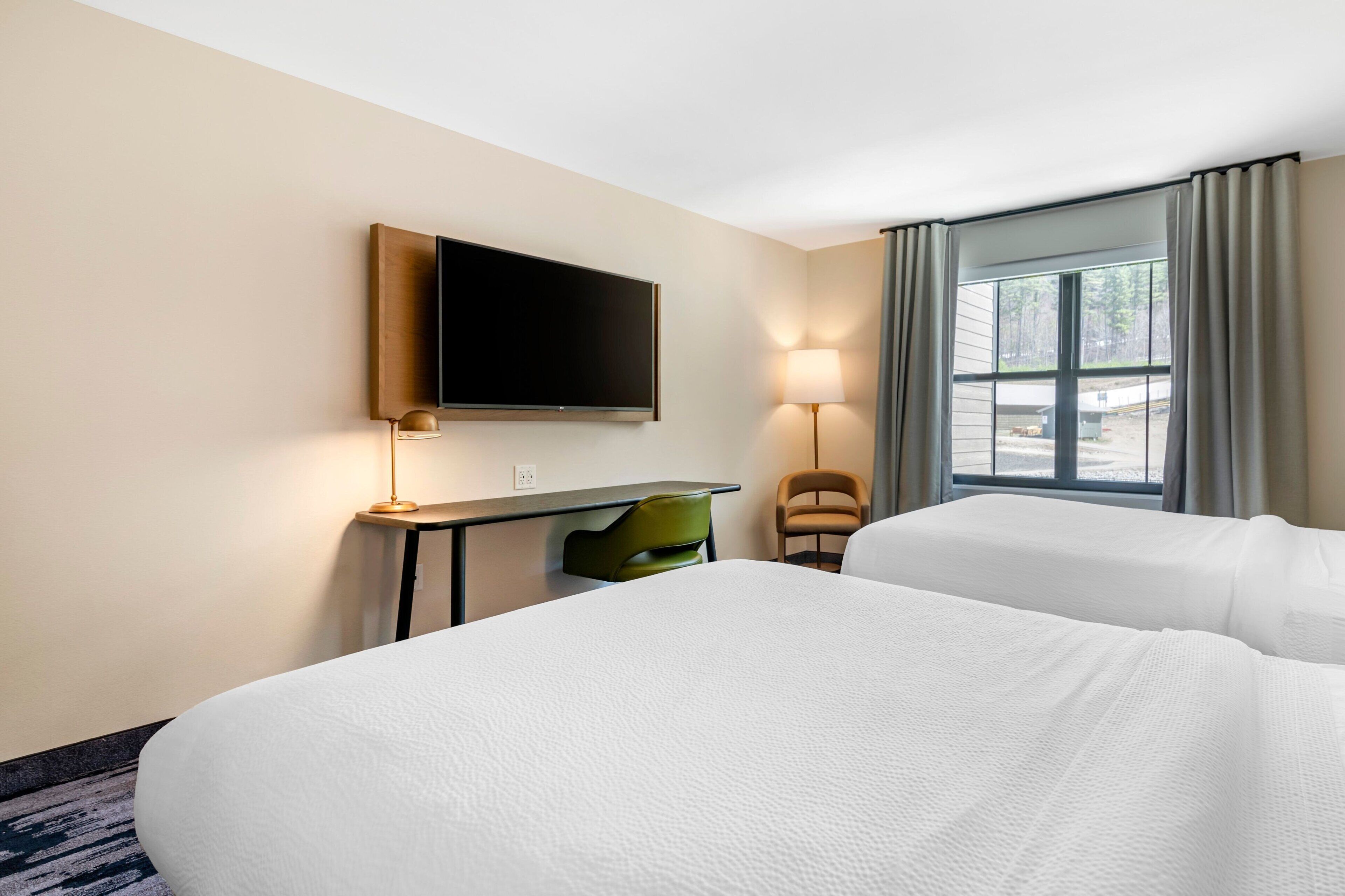 A hotel room with two beds, a flat-screen TV mounted on the wall, a desk with a lamp, and a chair.