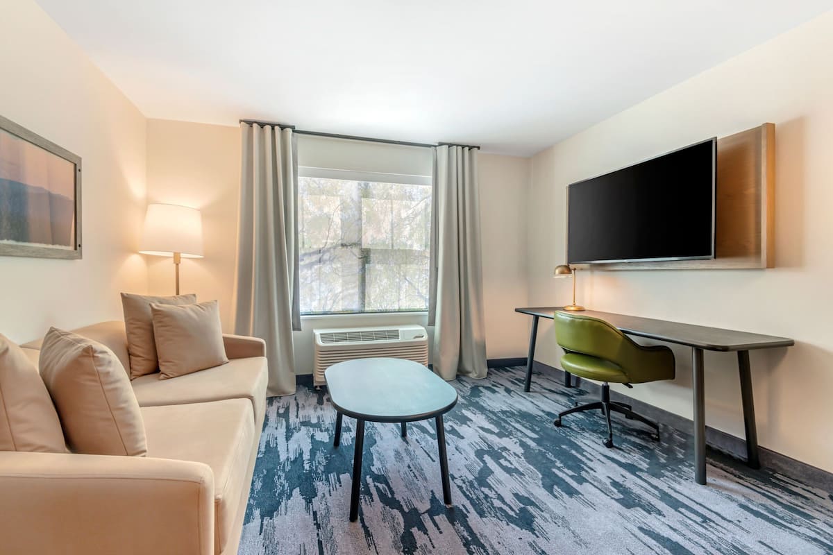 Habitación de hotel con sofá, una mesita, escritorio, silla, televisor y ventana con cortinas.
