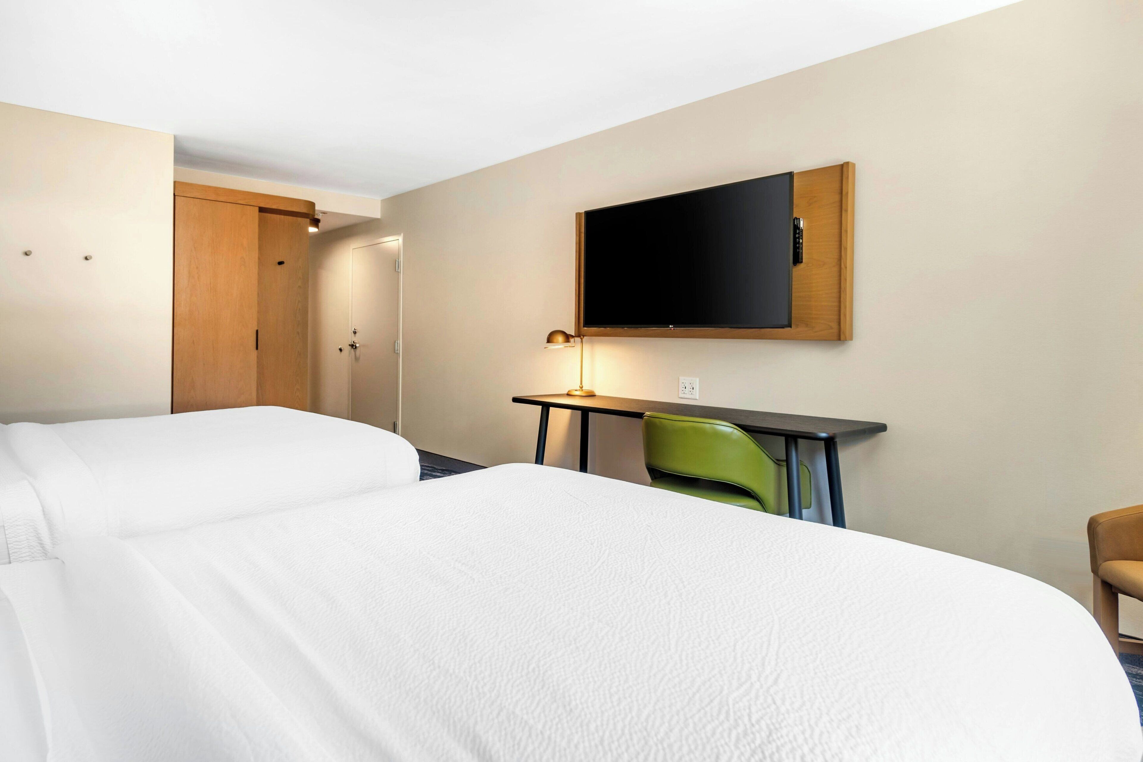 A hotel room with two beds, a desk with a lamp, a TV mounted on the wall, and a green chair.