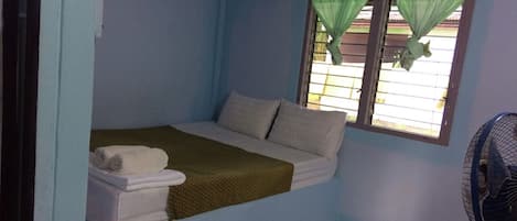 Una habitación pequeña con cama individual, una ventana cubierta por una cortina verde, una colcha blanca y una toalla blanca sobre la cama.