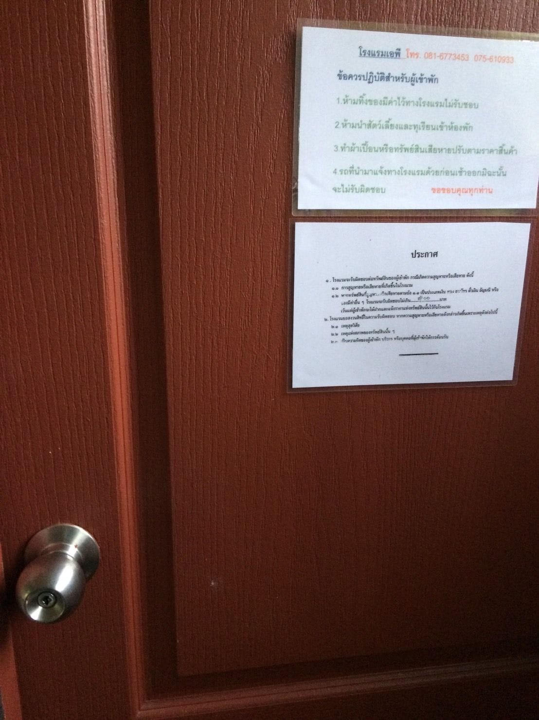 A wooden door with a metal handle and two white signs with Thai text.