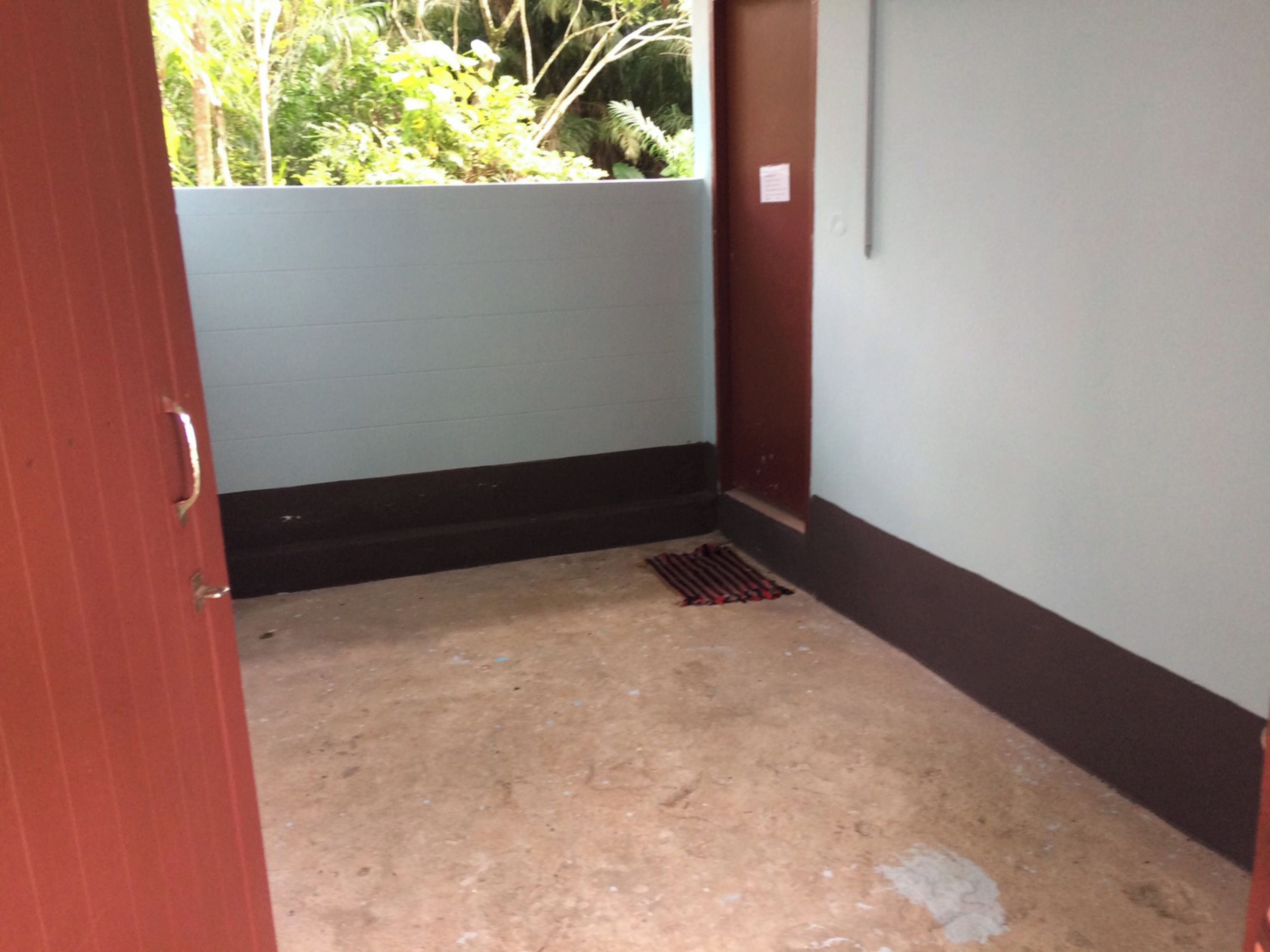 A narrow hallway with a concrete floor, a red door, and a window overlooking greenery.