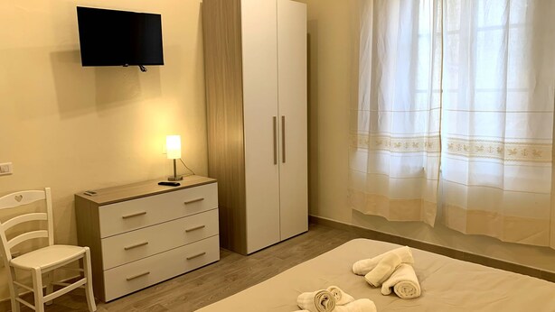 A bedroom with a bed, a TV mounted on the wall, a wardrobe, a chair, and a window with curtains. - Domo Achenza (Terralba)