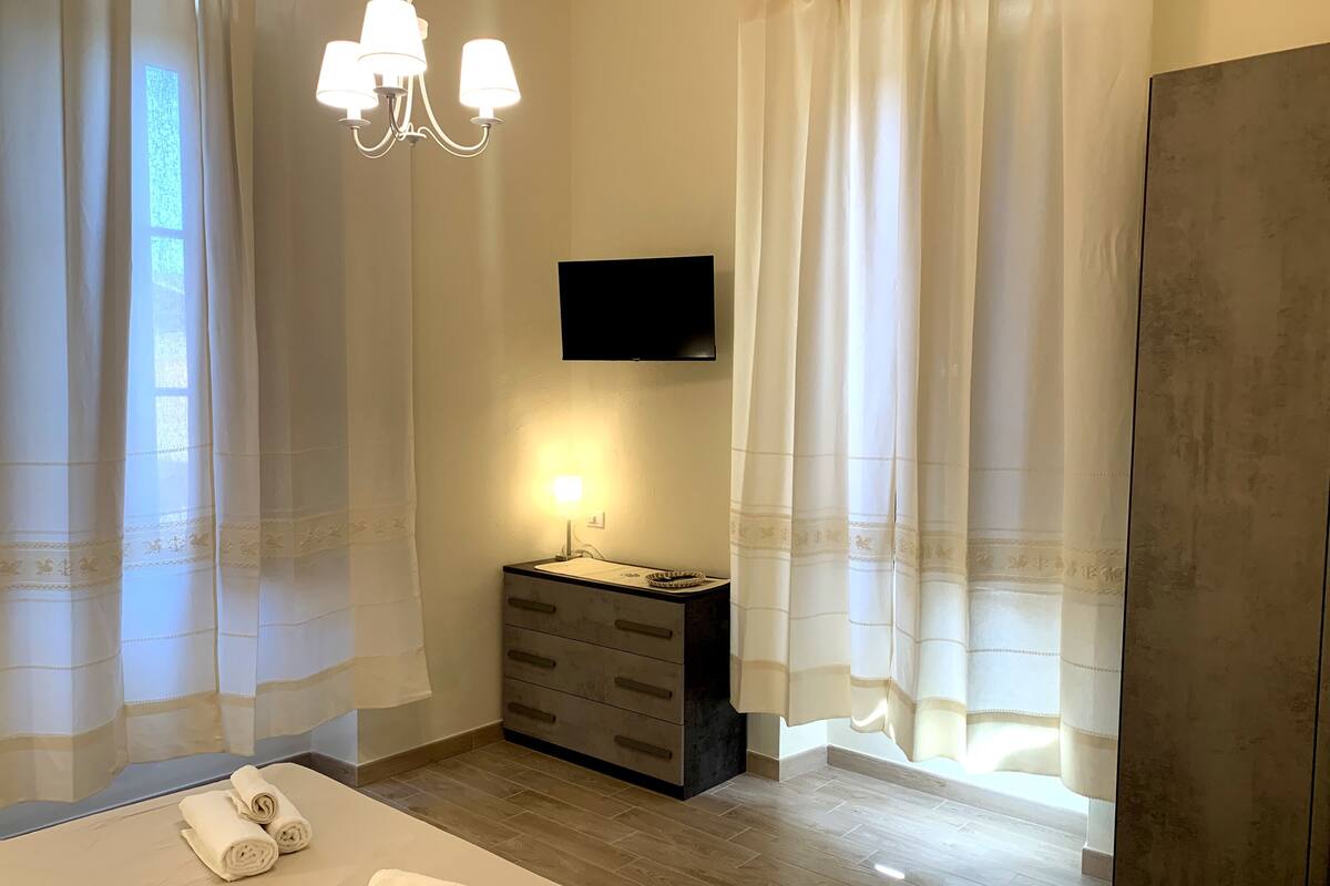 A hotel room with a bed, a bedside table with a lamp, a TV mounted on the wall, and a large window with curtains.