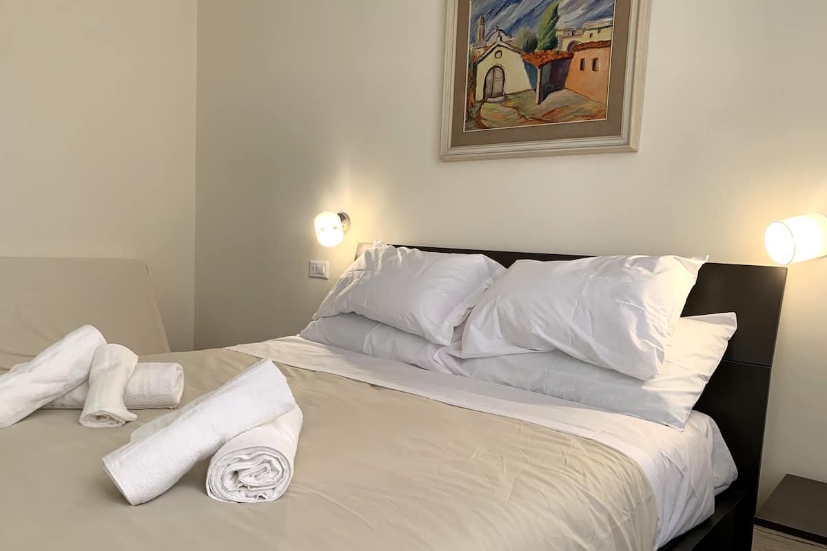 A neatly made bed with white linens and towels, a painting on the wall, and two wall-mounted light fixtures.