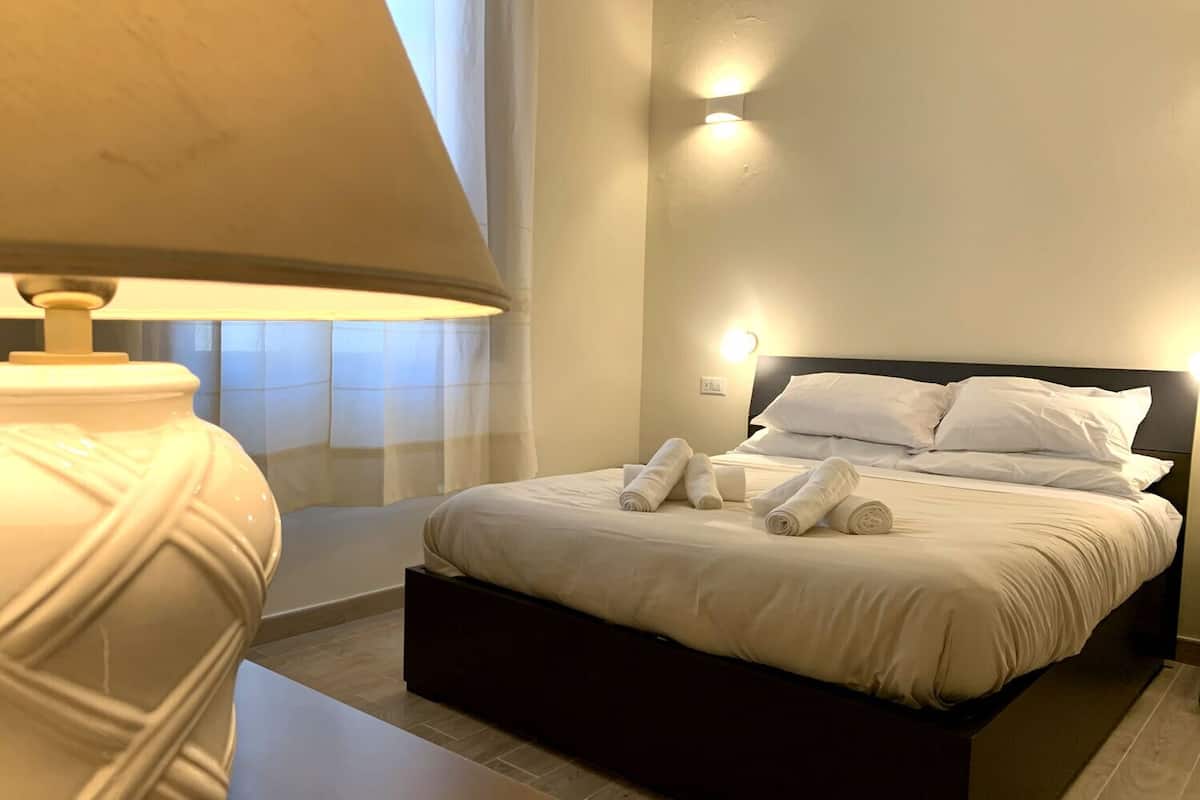 A neatly made bed with white linens and towels, a bedside lamp, and a wall-mounted light fixture.