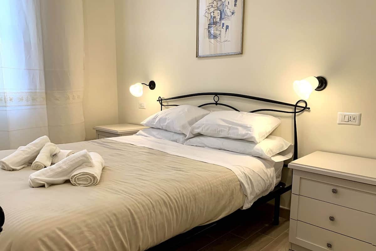 A neatly made bed with white linens and pillows, two bedside tables with lamps, a wall-mounted painting, and a power outlet on the wall.