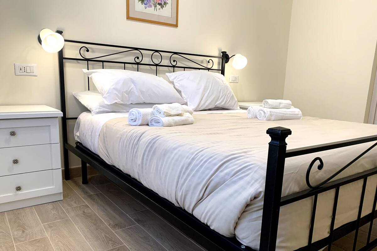 A neatly made bed with white linens and rolled towels, a white bedside table with drawers, and a black metal headboard.