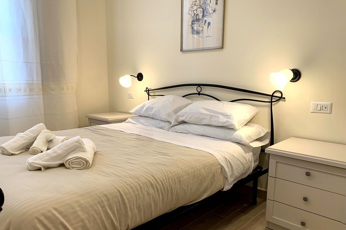 A neatly made bed with white linens and pillows, two bedside tables with lamps, a wall-mounted painting, and a power outlet on the wall.