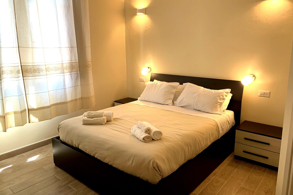 A neatly made bed with white linens and towels, two bedside tables, and wall-mounted lighting.