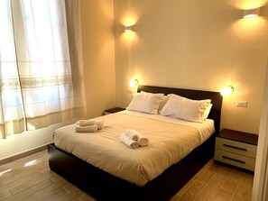 A neatly made bed with white linens and towels, two bedside tables, and wall-mounted lighting. - Domo Achenza (Terralba)
