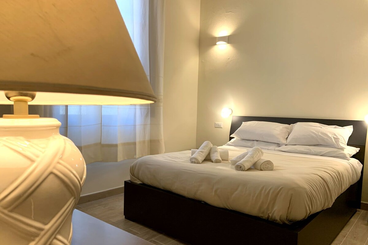 Un lit bien fait, avec du linge de lit et des serviettes blanches, une lampe de chevet et un luminaire fixé au mur.