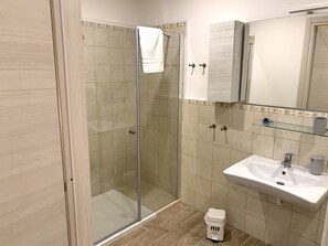 A bathroom with a glass shower enclosure, a sink, a mirror, and a small trash bin. - Domo Achenza (Terralba)