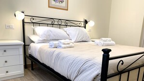 A neatly made bed with white linens and rolled towels, a white bedside table with drawers, and a black metal headboard. - Domo Achenza (Terralba)