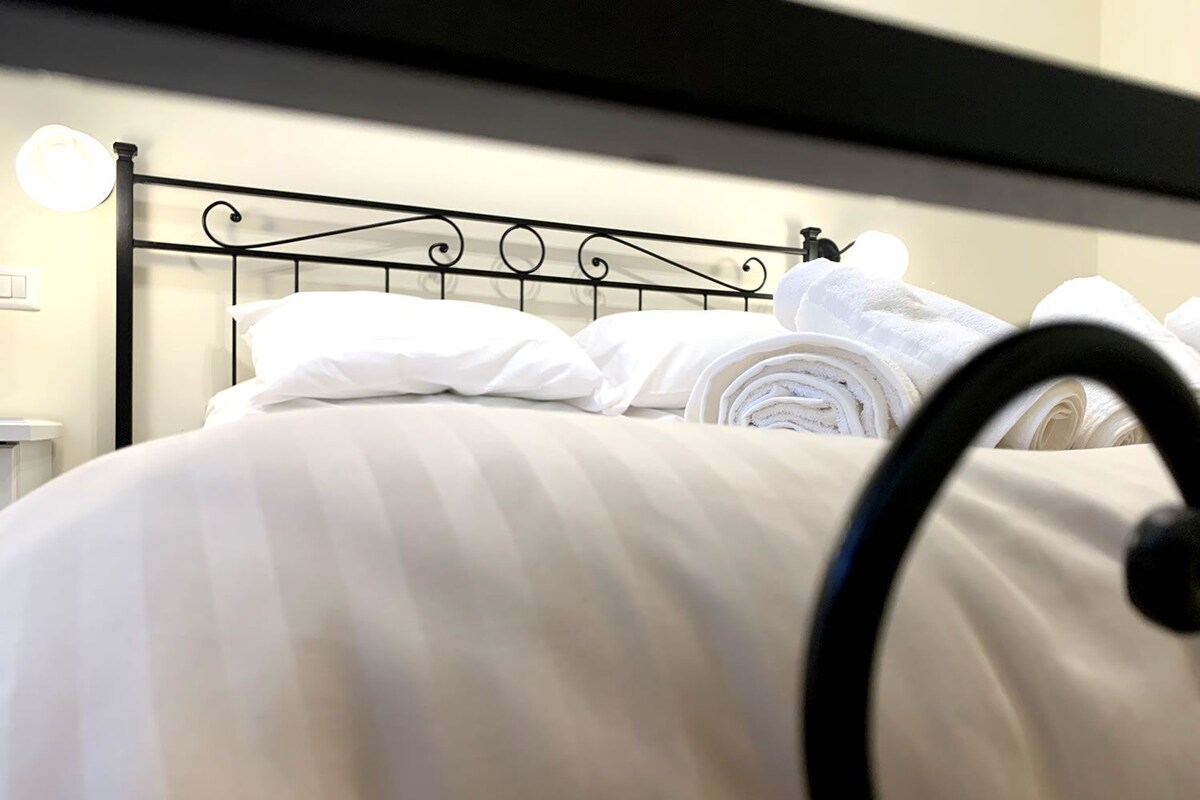 A neatly made bed with white linens and rolled towels, a black metal headboard, and bedside lamps.
